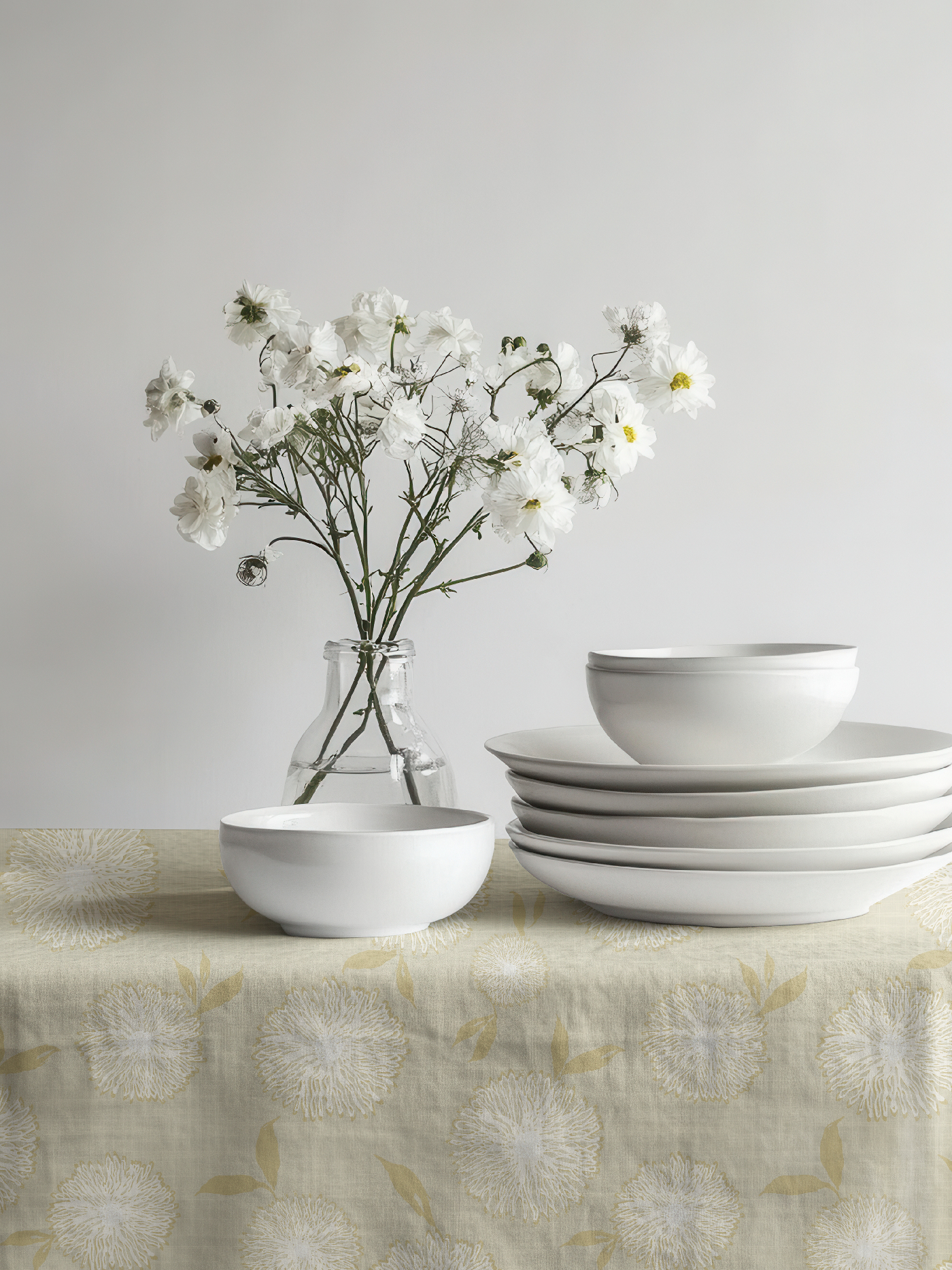 White ceramic dishes displayed on a table covered with the Dahlia Fields floral pattern from the Botanical Reverie collection in the Heritage Garden colorway, featuring delicate pale green flowers and foliage.